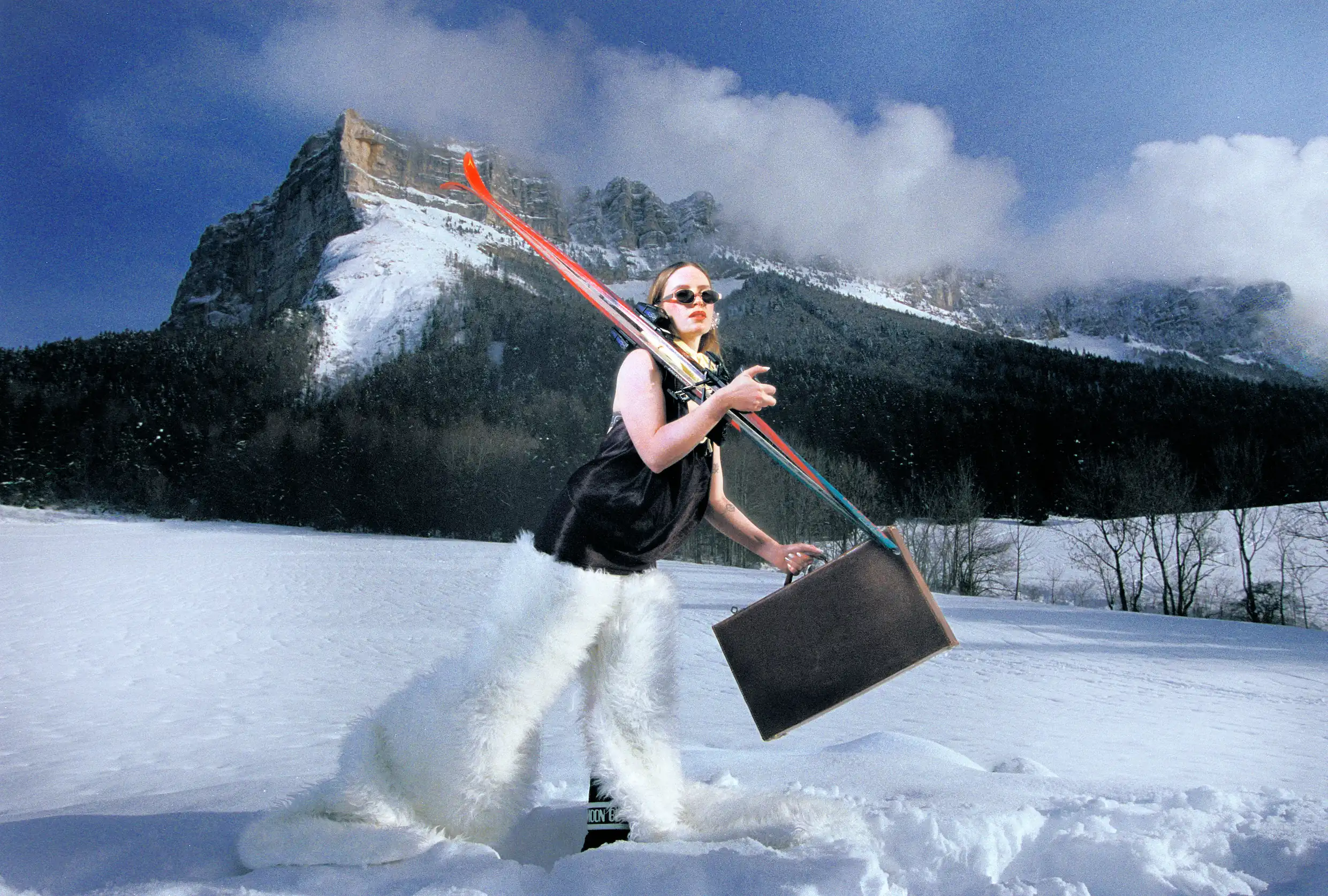 Wide frame of Marine D. crossing a snowfield with skis on her shoulder and a suitcase in hand
