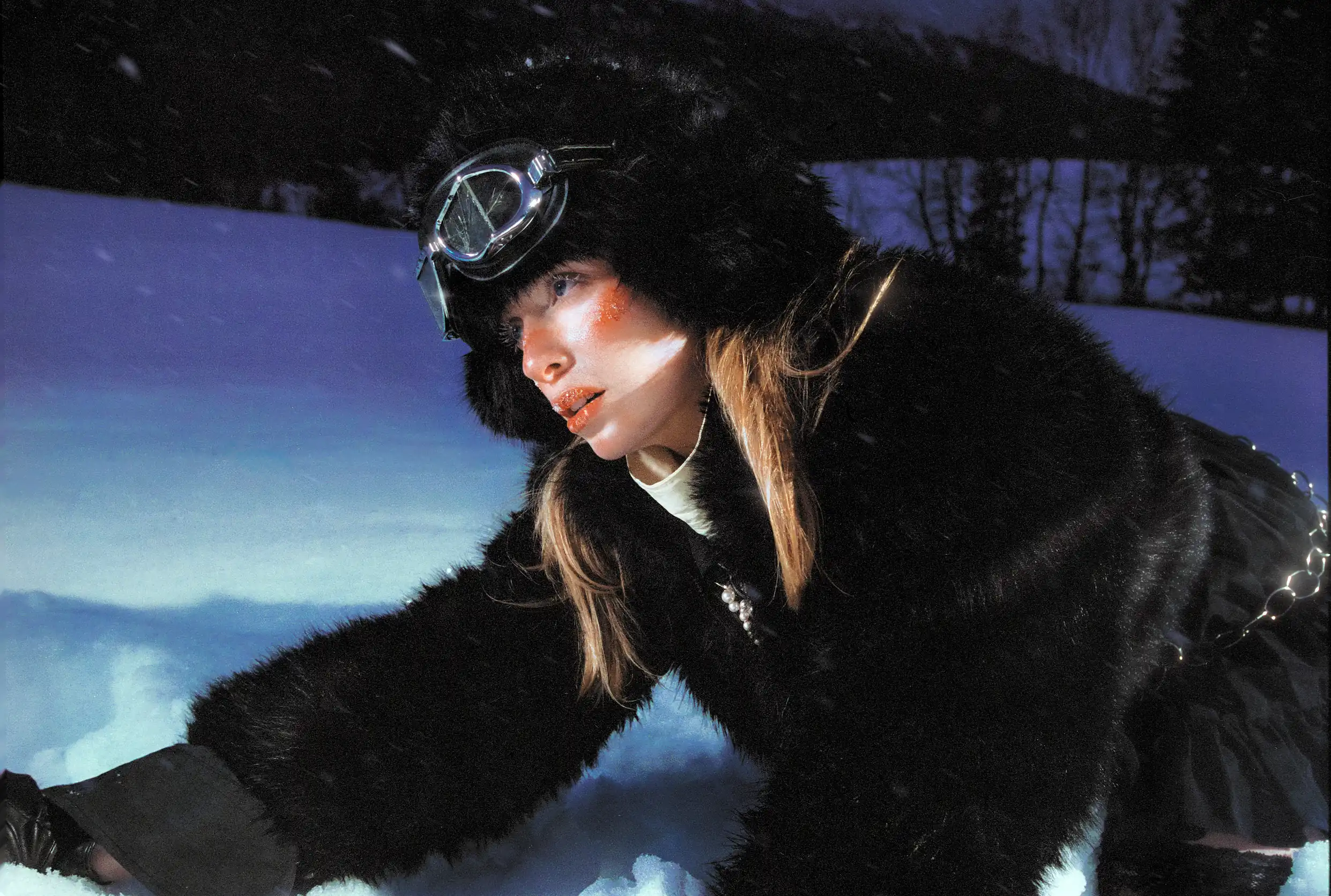 Marine D. crouched low over the snow in a black fur coat and ski goggles as flakes fall at dusk