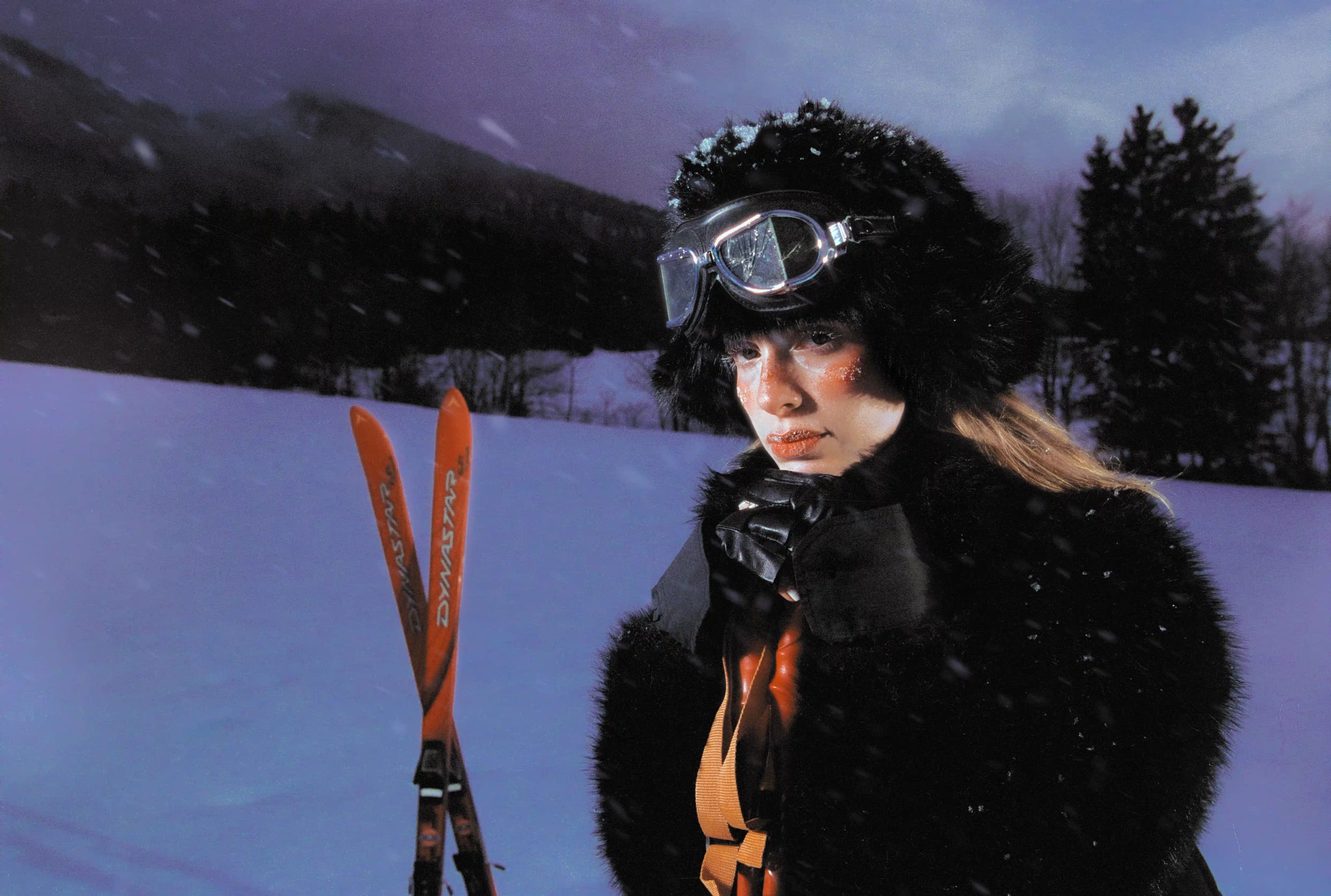 Marine D. facing the camera in falling snow, fur hat and goggles on, skis planted in the drift behind her