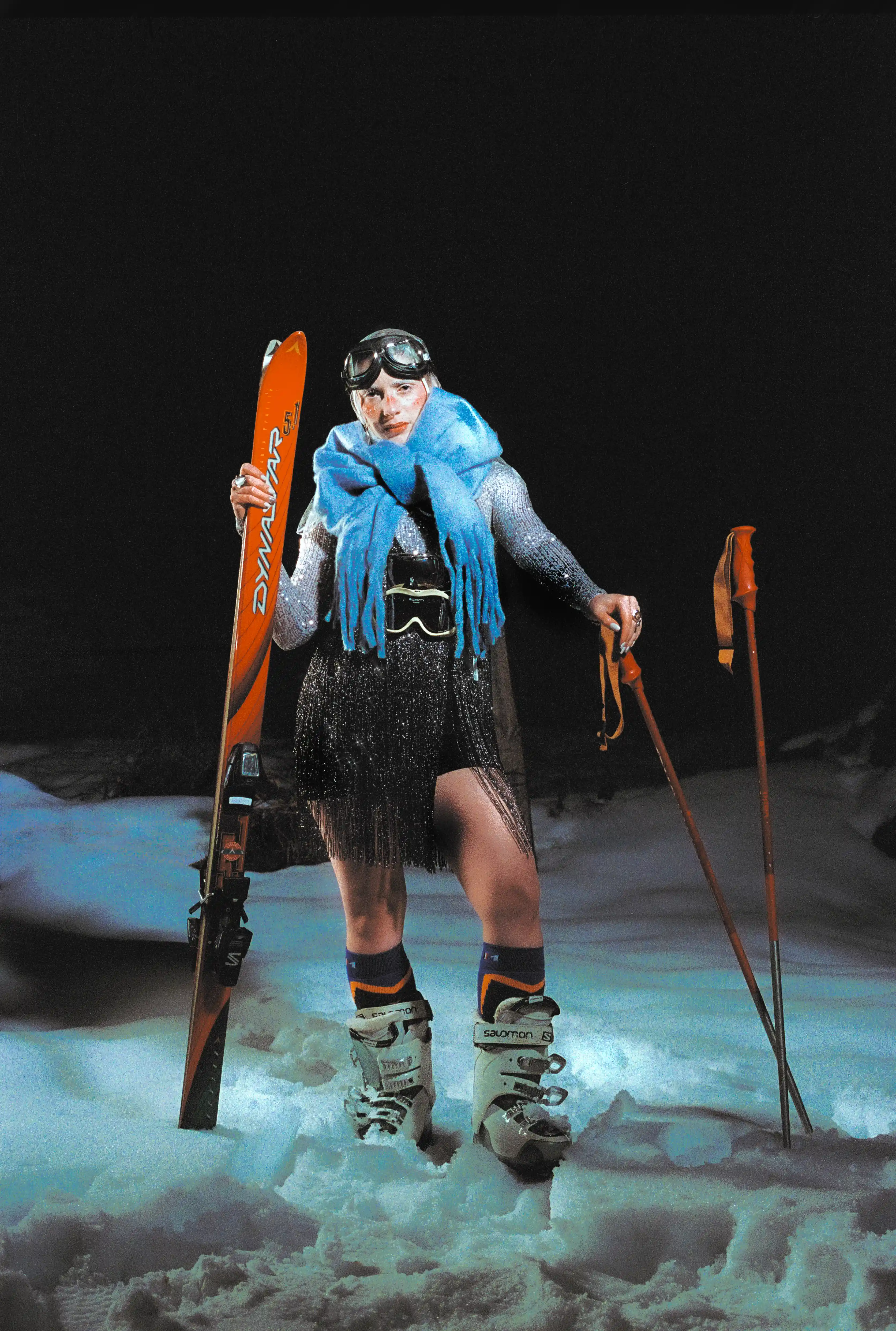 Full-length night portrait of Marine D. holding skis in one hand and poles in the other, blue knit scarf over a mesh dress
