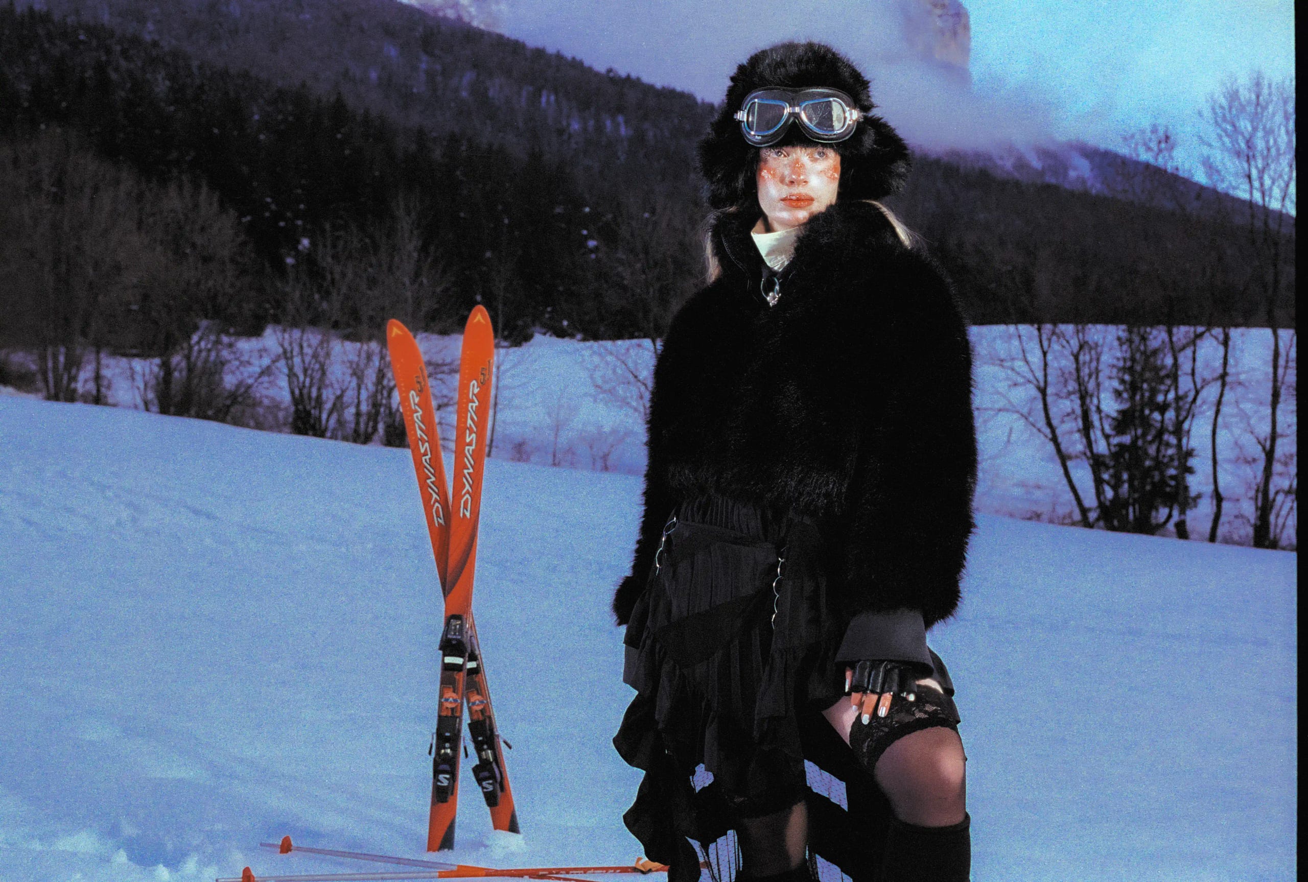 Marine D. standing in a black fur coat beside her planted skis, dark forest and mountain ridge behind