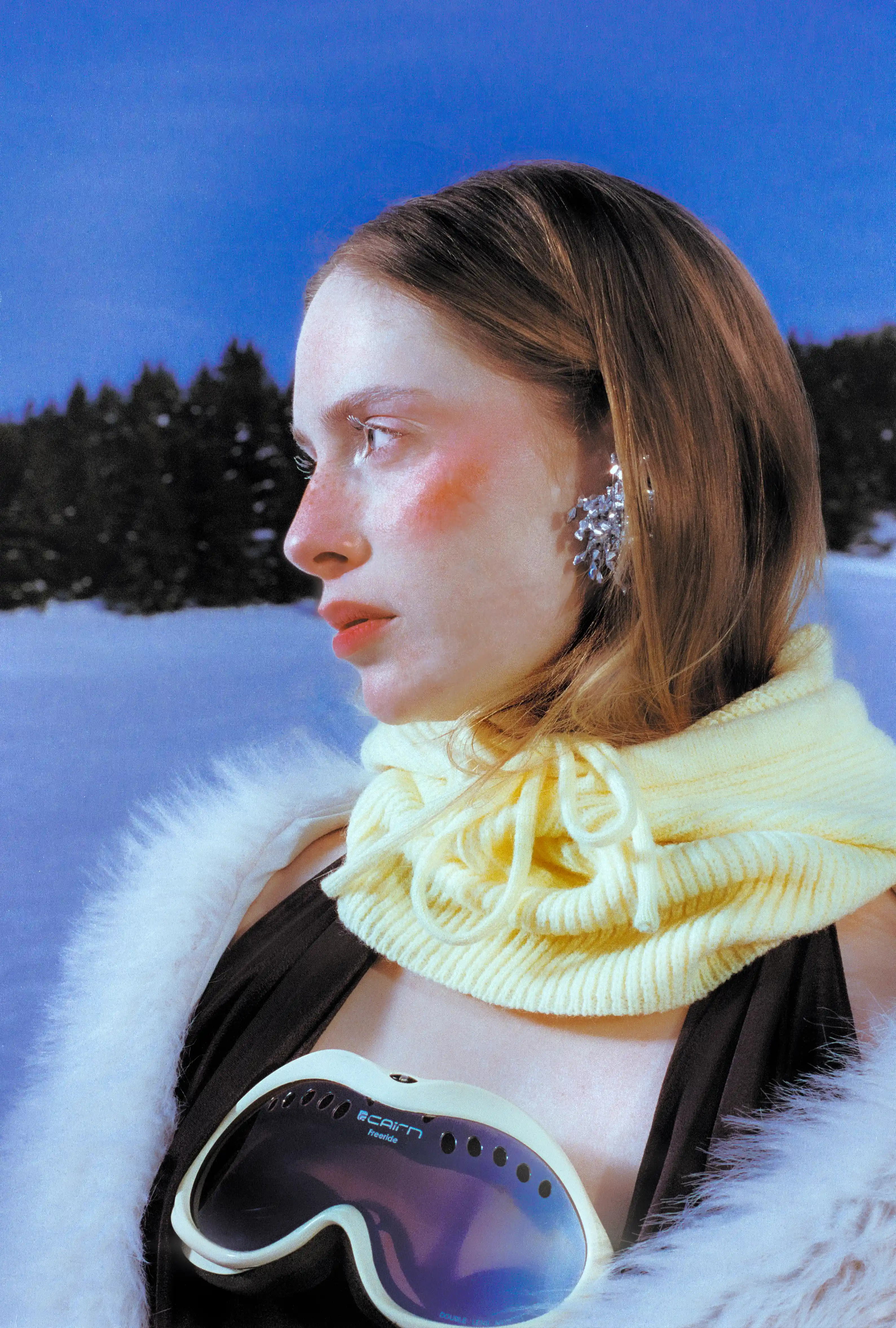 Profile of Marine D. in a yellow knit snood, ski goggles hanging at her chest against the pine line