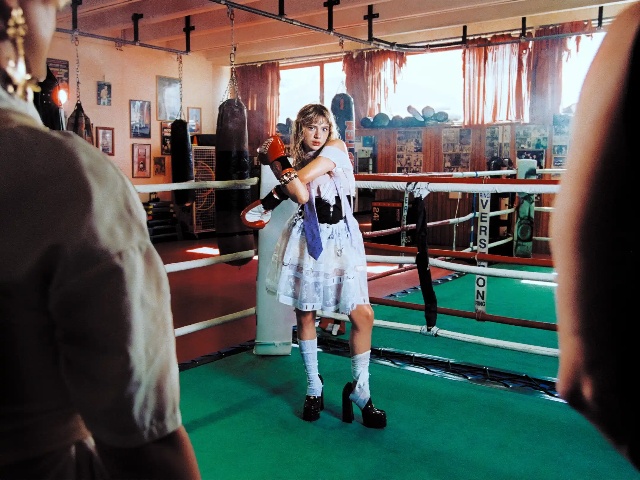 Full view of the boxing club past two out-of-focus figures, Romane Parc standing alone in the ring