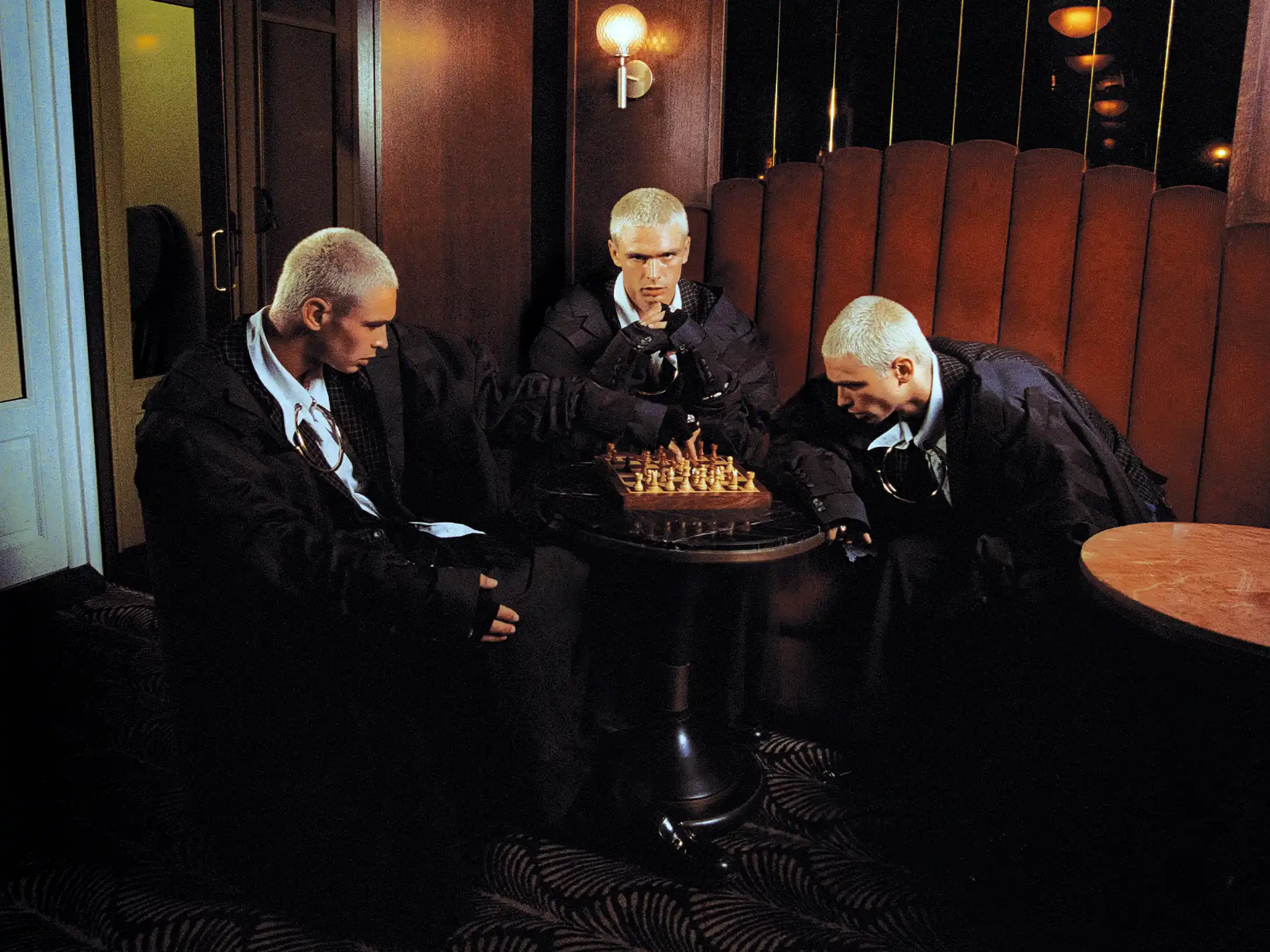 Three duplicates of Matheo gathered around a single chess table in a wood-panelled lounge