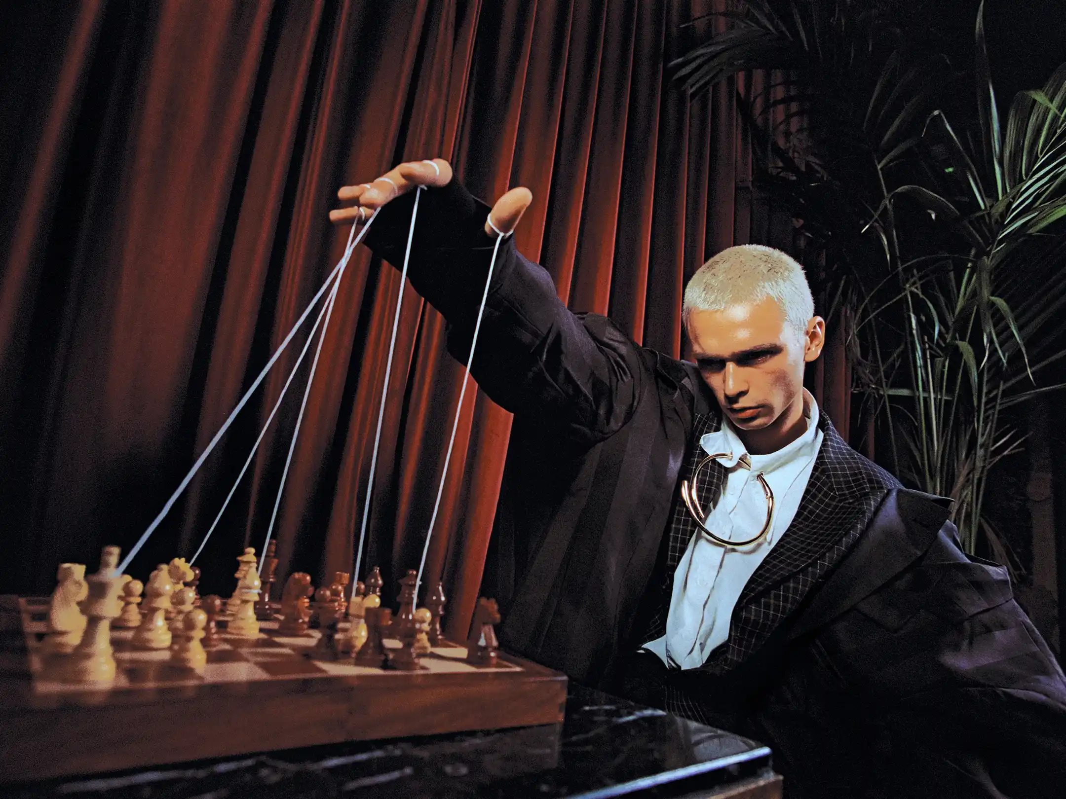 Matheo leaning over a pocket chessboard at a marble café table, wall sconce glowing behind him