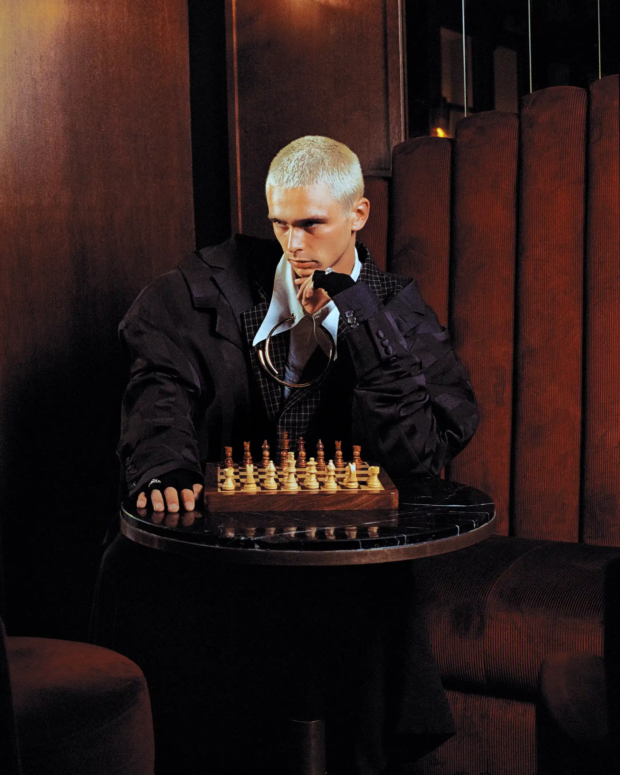 Matheo seated behind a full chess set, chin resting on a gloved hand, metal collar over a white shirt