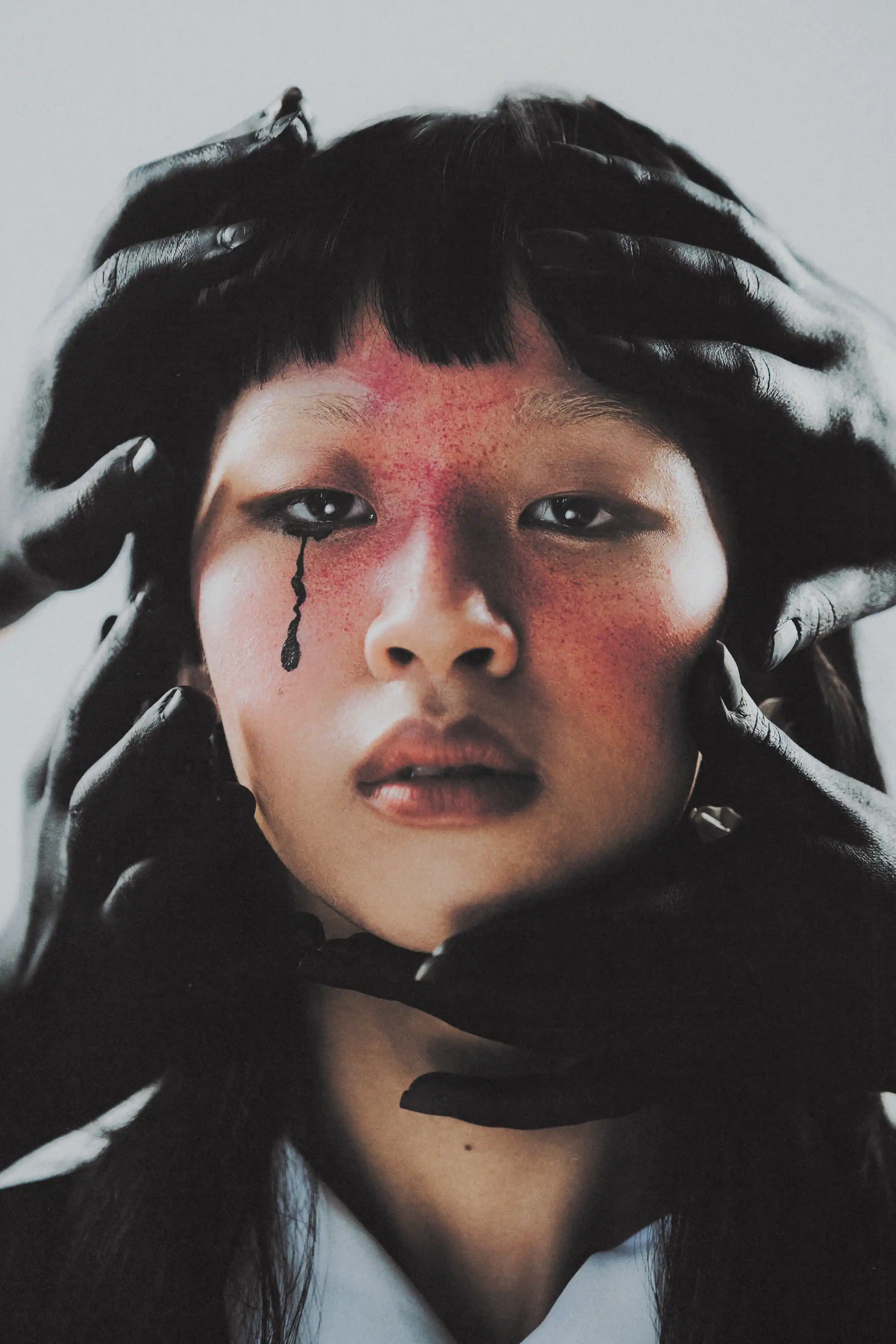 Karine P. facing the camera with red-flushed makeup and a painted black tear as gloved hands close around her head