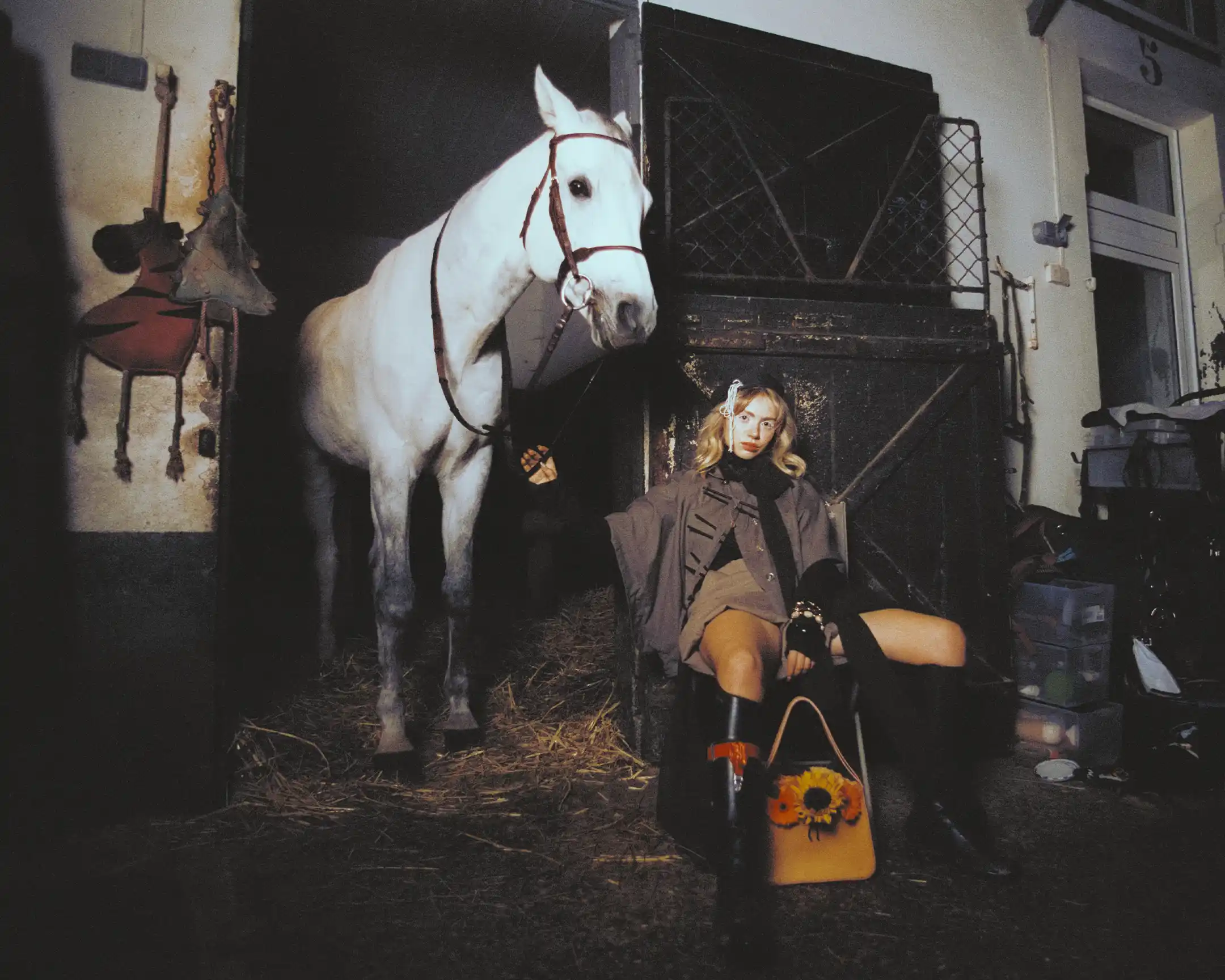 Wide stable frame: Cassalo standing on straw beside Romane Parc, seated in boots with a flower-trimmed bag at her feet