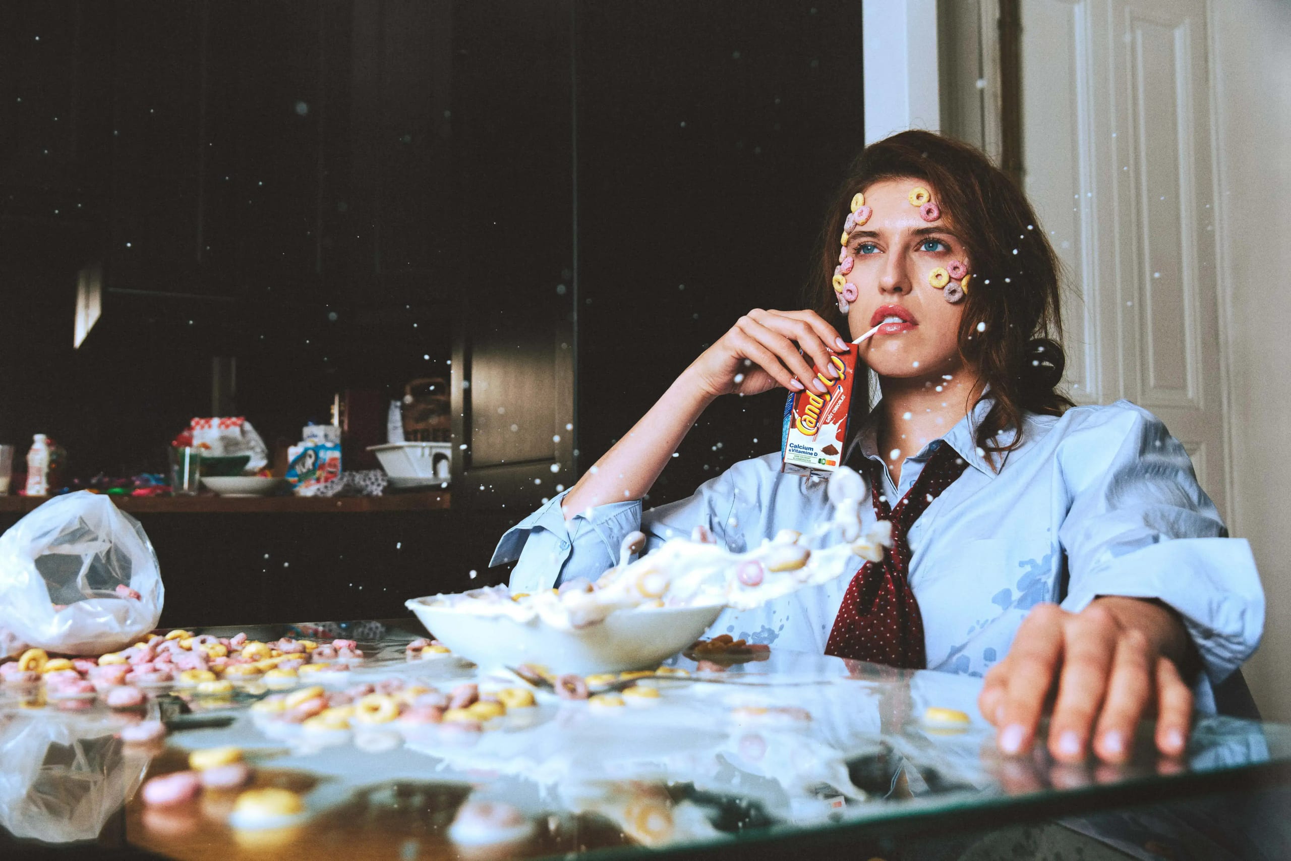 Model at a glass table flooded with milk and cereal, loops stuck to her face and a sweet packet in hand