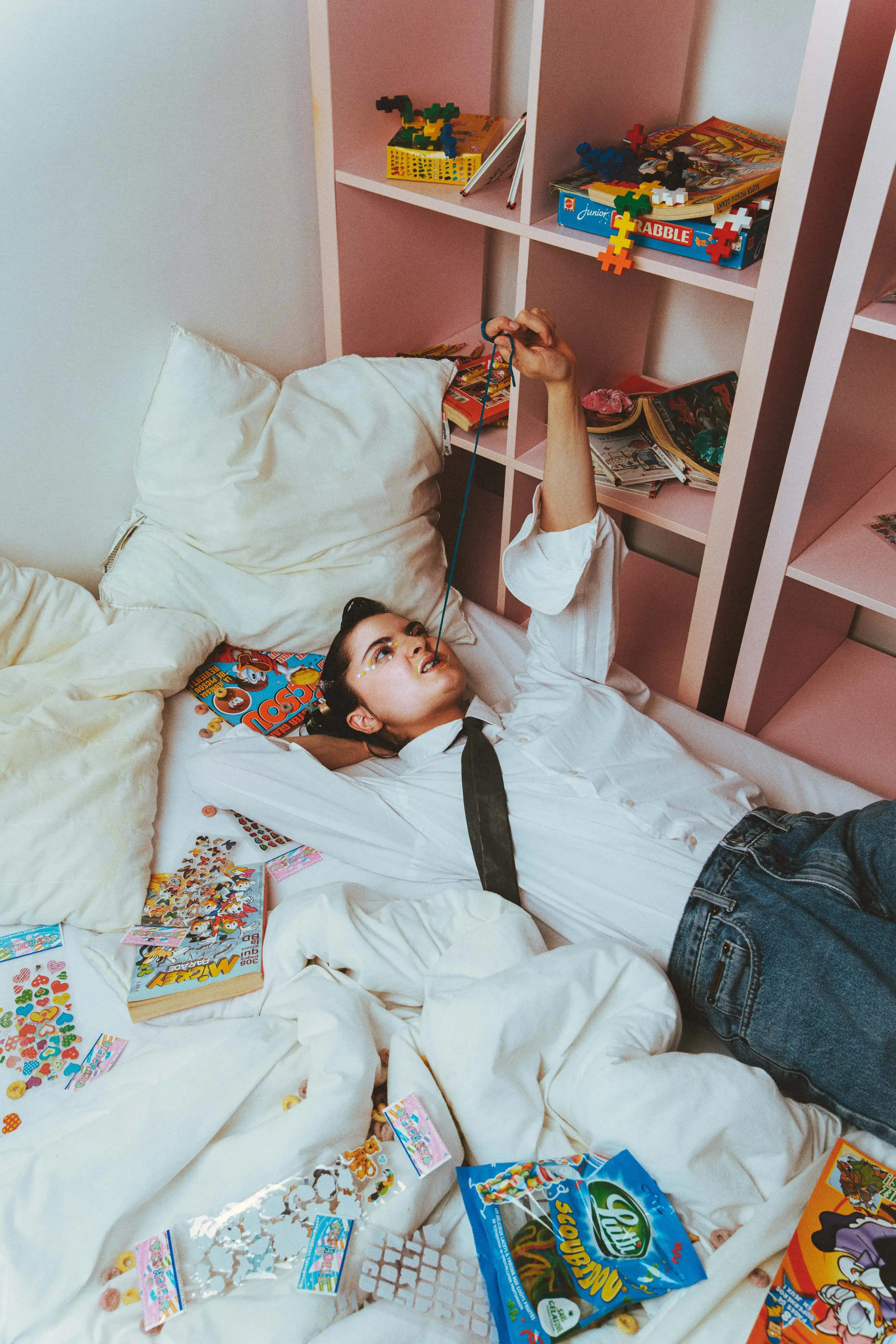 Model lying on a rumpled white bed strewn with comics, stickers and sweet wrappers, pink shelves of toys behind