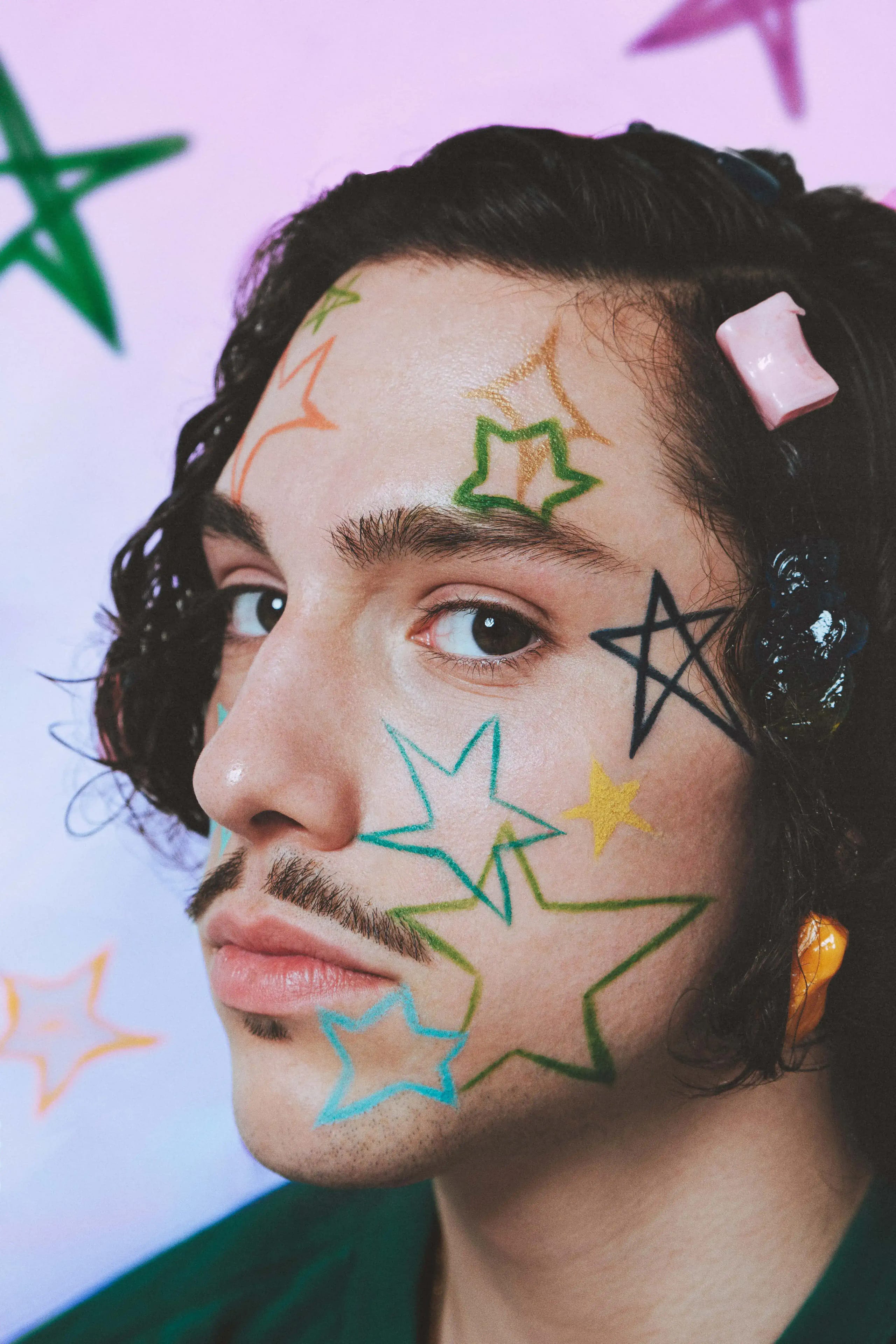 Close-up portrait with coloured stars drawn across the face and plastic clips in the hair, against a pastel painted backdrop