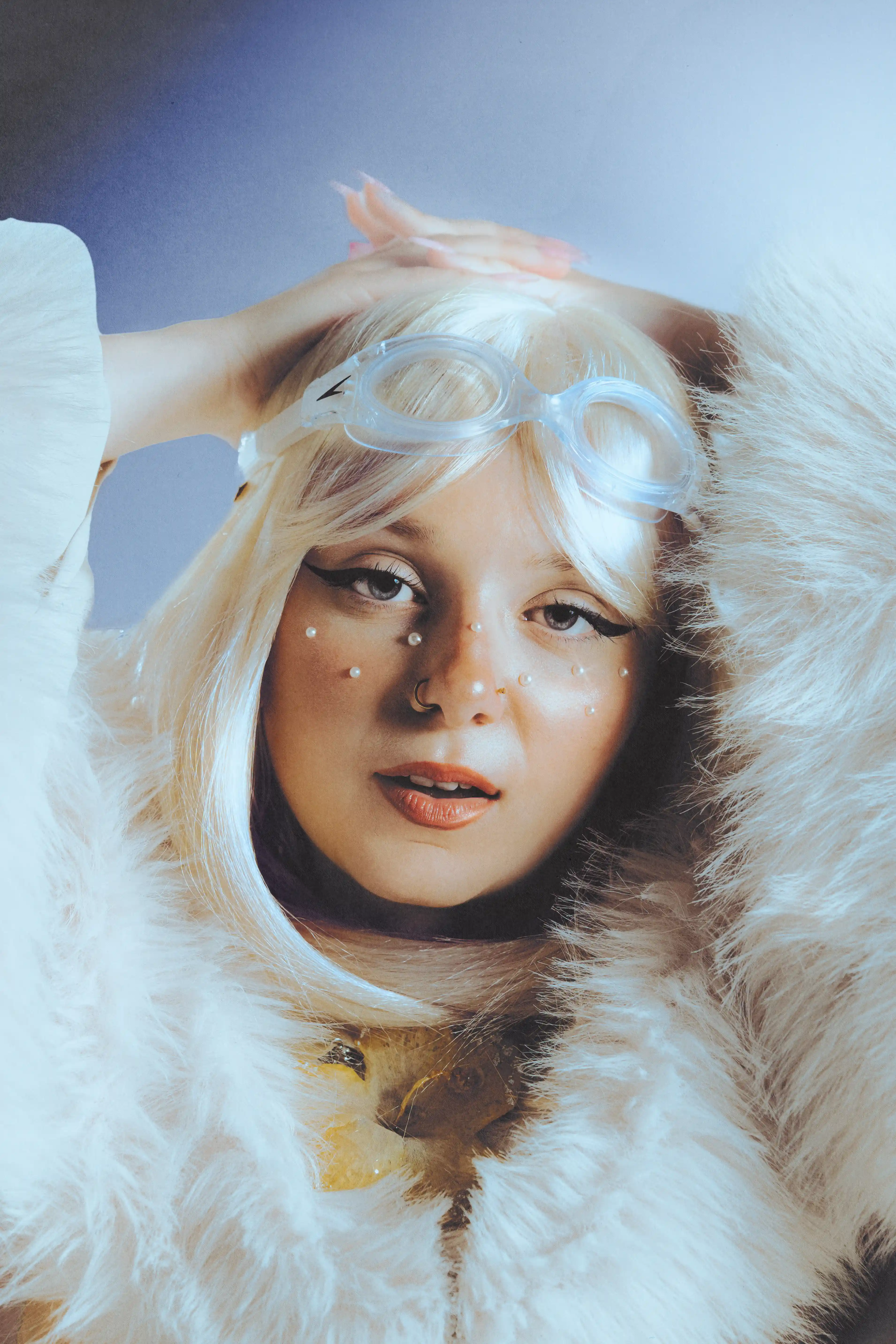 Close portrait of Julie in a white fur hood, clear goggles pushed onto her platinum wig and pearls set across her cheeks