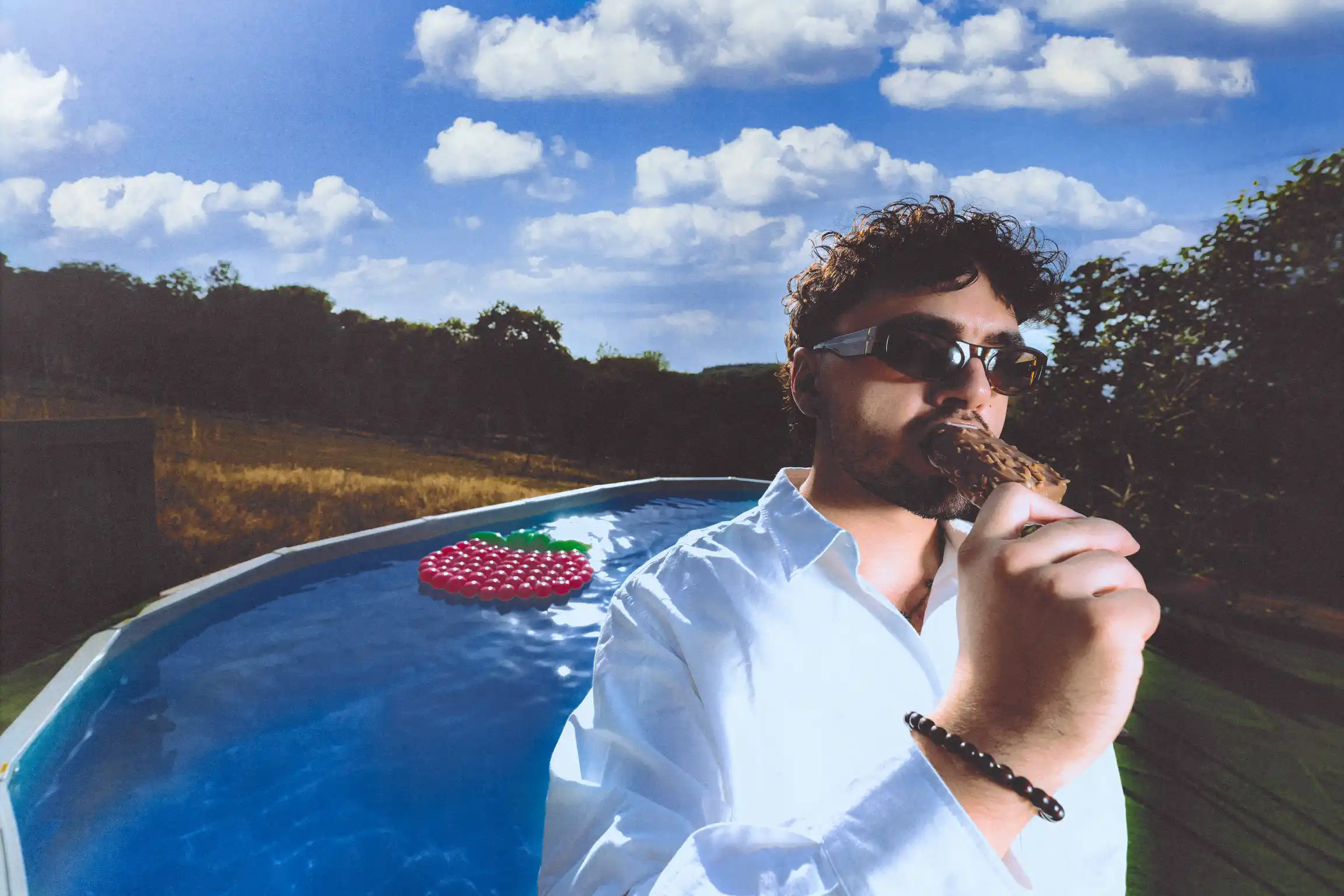 Low-angle portrait of Tyron B. biting into an ice cream bar, open white shirt and slim sunglasses against the water