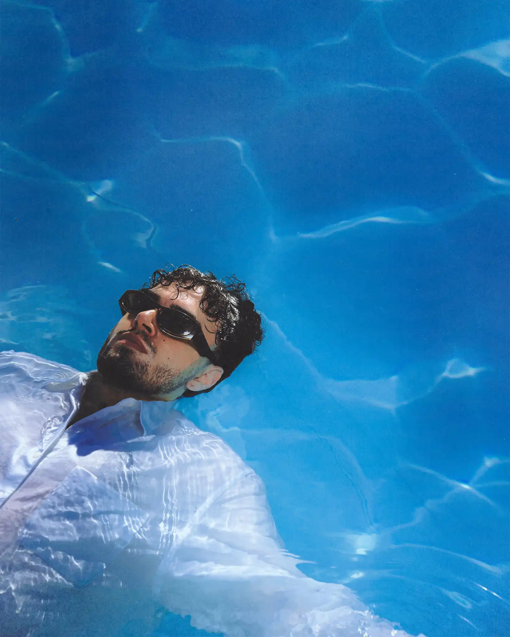 Overhead frame of Tyron B. floating on his back in soaked clothes, face breaking the blue surface