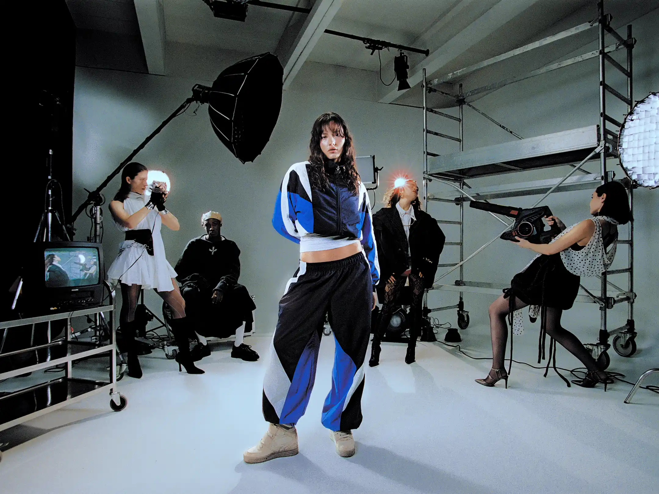 Wide studio frame of Taabzzi posing on the white set while the whole cast shoots her from every side