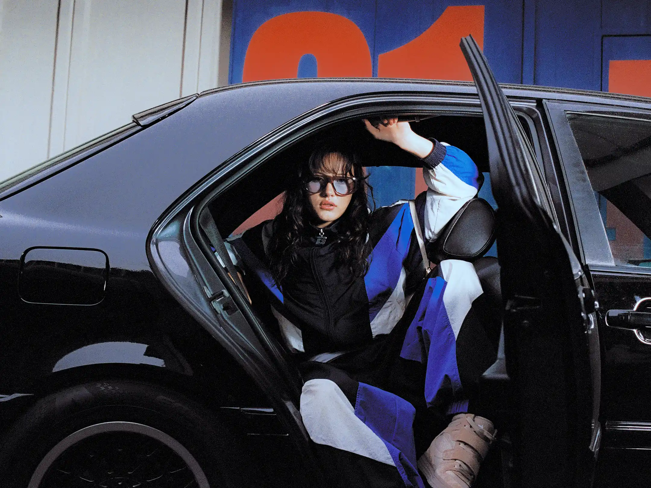 Taabzzi holding the open door of a black car in oversized sunglasses, painted signage filling the wall behind
