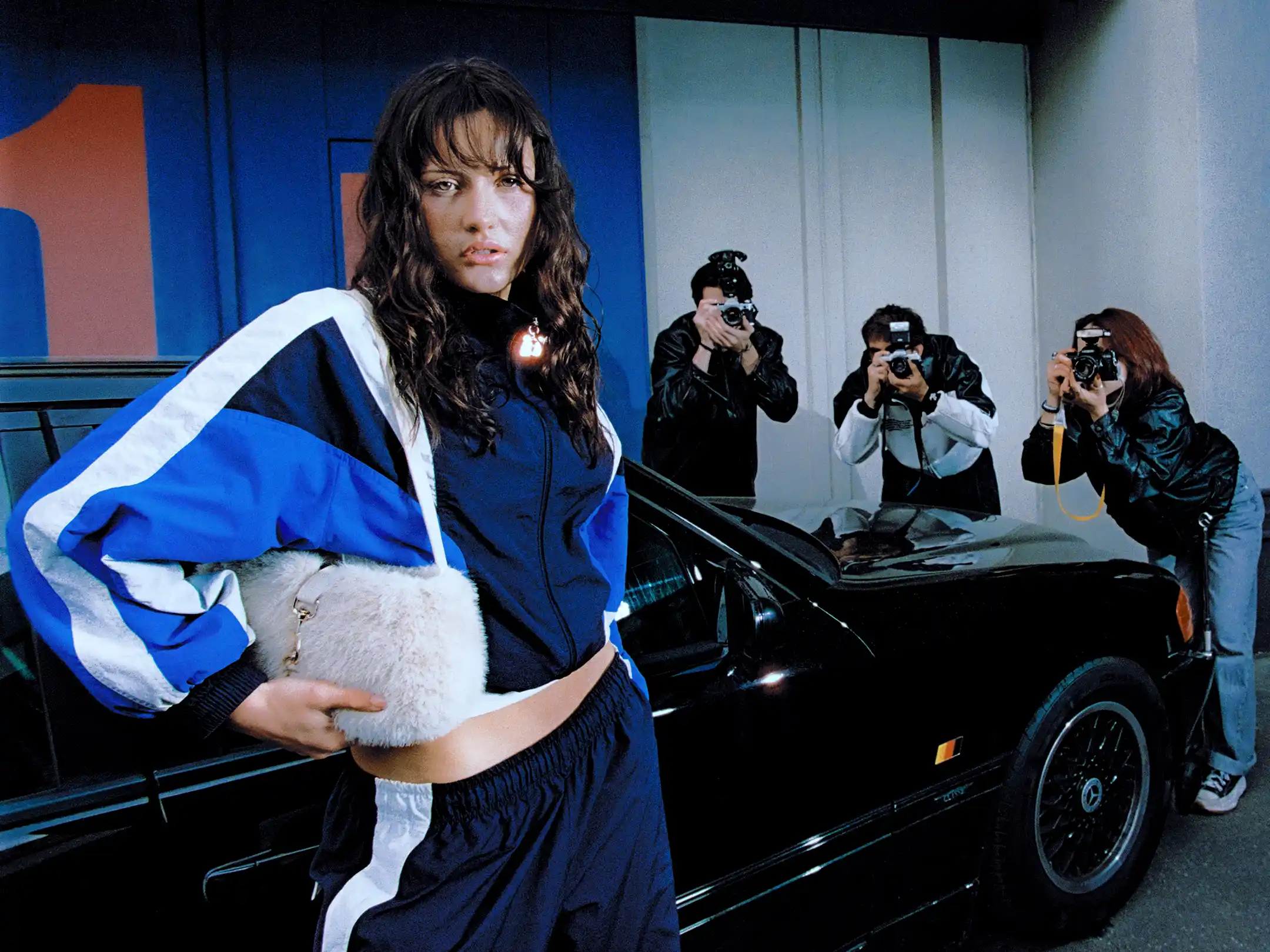 Taabzzi leaning against a black car with a fur bag as three photographers shoot her from behind