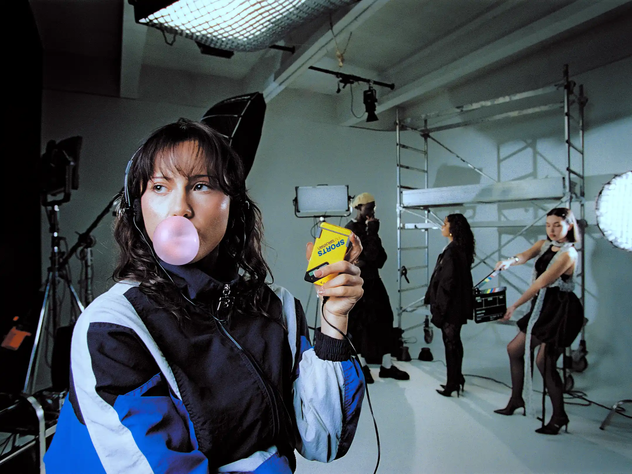 Taabzzi blowing a bubble while holding up a boxed roll of film, the crew still working on set behind her